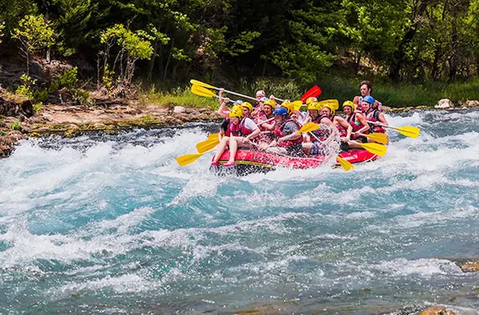 Multiple groups of people in colorful inflatable rafts navigate a brown river surrounded by dense green jungle vegetation. Participants wear life jackets and helmets, paddling through the water with lush forest and tall grass visible along the riverbanks. The scene conveys adventure and excitement of white water rafting in a tropical environment.